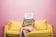 © LIGHTFIELD STUDIOS - asian woman holding newspaper with fake news in front of face while sitting on yellow sofa isolated on pink