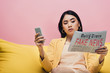 © LIGHTFIELD STUDIOS - serious asian woman holding newspaper with fake news and smartphone on yellow sofa isolated on pink