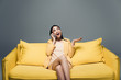 © LIGHTFIELD STUDIOS - happy asian businesswoman sitting on yellow sofa with crossed legs and talking on smartphone on grey background