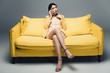 © LIGHTFIELD STUDIOS - pensive asian businesswoman sitting on yellow sofa with crossed legs and talking on smartphone on grey background