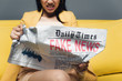 © LIGHTFIELD STUDIOS - cropped view of angry asian businesswoman sitting on yellow sofa and reading newspaper with fake news on grey background