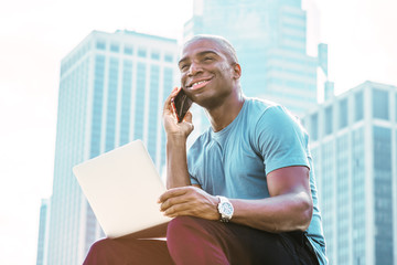 Wall Mural - Young African American Businessman traveling, working in New York, wearing blue T shirt, sitting in front of business district, working on laptop computer, talking on cell phone, looking up, smiling..