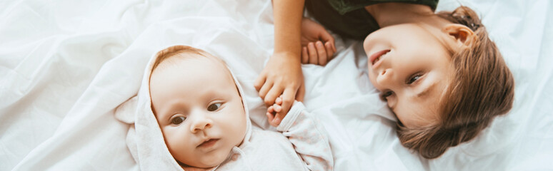 Wall Mural - panoramic shot of smiling child holding hand of little sister lying on white bedding