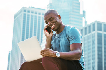 Wall Mural - Young African American Businessman traveling, working in New York, wearing blue T shirt, sitting in front of business district, looking down, working on laptop computer, talking on cell phone, smiling