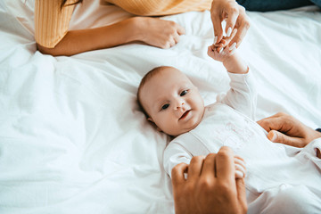 Wall Mural - cropped view of mother and dad playing with smiling baby lying on white bedding