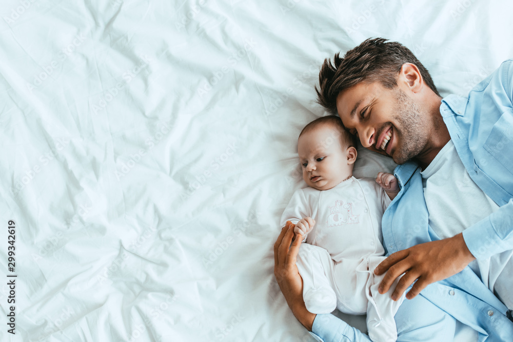 top view of happy father hugging adorable baby while lying on white bedding - obrazy, fototapety, plakaty 