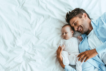 Wall Mural - top view of happy father hugging adorable baby while lying on white bedding