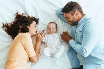 Wall Mural - happy mom and dad gently touching adorable baby while lying on white bedding together