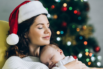 Wall Mural - happy woman in santa hat sitting with closed eyes and holding sleeping baby