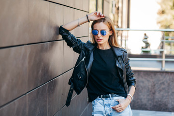 Wall Mural - Girl wearing black t-shirt, glasses and leather jacket posing against street , urban clothing style. Street photography