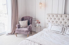 White Classic Bedroom Interior With New Year's Holiday Bouquet In A Vase, A Gently Pink Gift Present Box On The Bedside Glass Table And Classic Armchair In Lavender Colors
