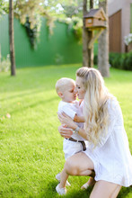 Young Caucasian Mother Sitting With Child On Grass In Yard. And Kissing Little Boy