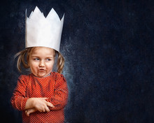 Little Girl With Paper Crown Standing With Arms Crossed And Scary Face Grimace. Portrait On Dark Blue Wall Background.