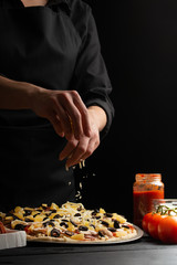 Wall Mural - Chef cooks Italian pizza, sprinkles with mazarella cheese. Freeze in motion. Against the background of pizza ingredients. Black background, pizzeria, recipe book,restaurant business. Vertical photo.