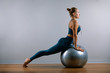 © Georgii - Young, beautiful, athletic girl doing exercises on fitball in the gym on a gray background. Sporting Slavic girl in a blue, green suit. Copy space, gray background, sport banner for advertising.