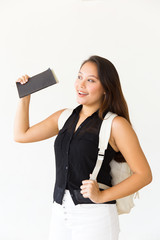 Wall Mural - Happy pensive student girl with notebooks waving hello, holding backpack strap. Young Latin woman standing isolated over white background. Communication concept