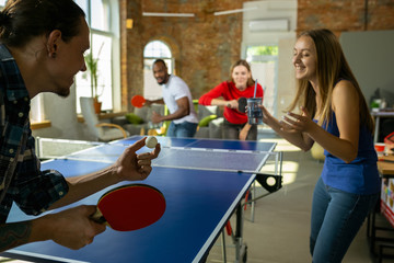 Wall Mural - Young people playing table tennis in workplace, having fun. Friends in casual clothes play ping pong together at sunny day. Concept of leisure activity, sport, friendship, teambuilding, teamwork.