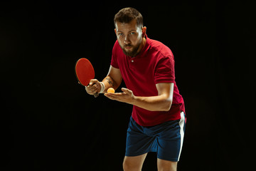 Young man plays table tennis on black studio background. Model in sportwear plays ping pong. Concept of leisure activity, sport, human emotions in gameplay, healthy lifestyle, motion, action, movement