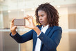 Focused female professional taking picture on cellphone outside. Young African American business woman standing near outdoor glass wall. Photo concept