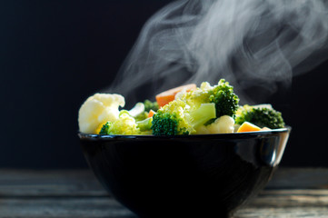 Wall Mural - Vegetables, I steaming in black bowl on woodeb tabke . Boiled hot Healthy food on table on black background. hot food concept.