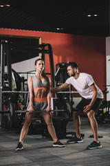 Wall Mural - handsome trainer supporting attractive sportswoman lifting barbell