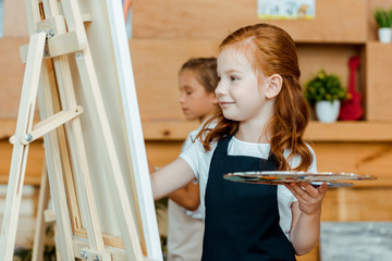 selective focus of cheerful redhead kid holding palette near easel and child in art school