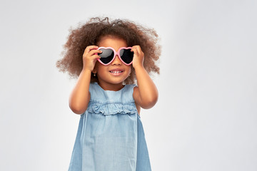 Wall Mural - childhood, summer accessory and valentines day concept - happy little african american girl in heart shaped sunglasses over grey background