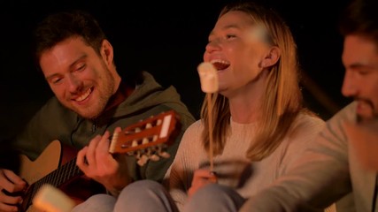 Poster - leisure and people concept - group of smiling friends sitting at camp fire on beach, roasting marshmallow and playing guitar at night