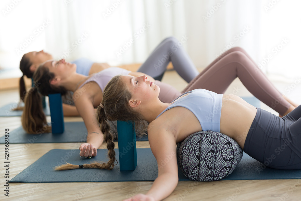 Restorative yoga with a bolster. Group of three young sporty attractive women in yoga studio, lying on bolster cushion, stretching and relaxing during restorative yoga. Healthy active lifestyle. - obrazy, fototapety, plakaty 