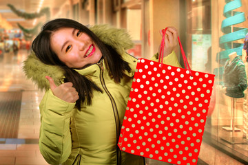 young happy and beautiful Asian Korean woman holding red shopping bag buying Christmas present in city mall smiling excited and cheerful in warm Winter hood coat