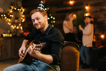 Wall Mural - Portrait of young man in the festive hat playing on the guitar against the background of talking friends. Christmas tree with garland and wall with festive illumination in background.