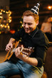 © dikushin - Portrait of young cheerful man in the festive hat playing on the guitar against the background of talking friends. Christmas tree with garland and wall with festive illumination in background.