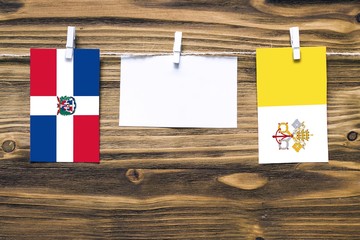 Hanging flags of Dominican Republic and Vatican City attached to rope with clothes pins with copy space on white note paper on wooden background.Diplomatic relations between countries.