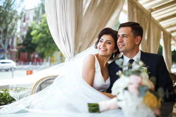 Young bride and groom in a cafe. Husband and wife hold each other's hands. Loving couple in a restaurant. Groom kissing a bride. Pretty bride and stylish groom. Wedding day. Marriage.