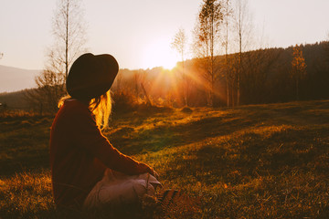 A dreamy young girl or woman traveler in a fedora hat sits in the grass and enjoys the mountain view, the warm rays of the autumn sun and a beautiful sunset.