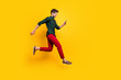 Full length profile side photo of positive cheerful guy use smartphone search discounts on social media jump run fast hurry wear green shirt red pants trousers isolated yellow color background