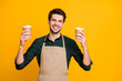 Photo of cheerful white positive toothy beaming barista hold green shirt two disposable paper cups of tea and coffee for you to choose isolated vibrant color background