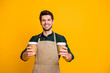 © deagreez - Portrait of positive cheerful guy worker in coffee bar shop hold paper card cup offer to his client mug with hot cappuccino wear apron green shirt isolated over yellow color background