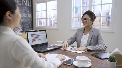 Wall Mural - female business people shaking hands. two asian chinese ladies cooperation partners handshaking in cafe bar. group of women workers in suit sitting at table with laptop and documents