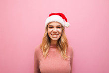 Portrait Of Smiling Girl In Christmas Hat On Pink Background, Looks Into Camera And Laughs. Joyful Lady Santa Is Smiling, Wearing A Pink Warm Sweater And Santa Hat. Christmas Concept