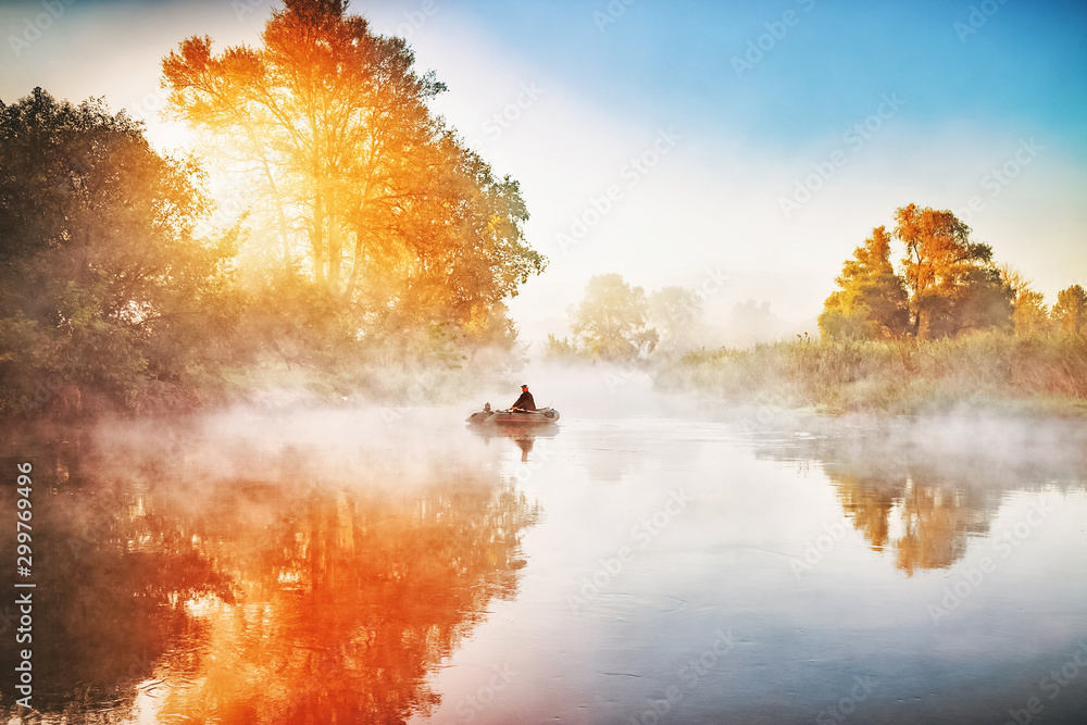 Fisherman in rubber boat making fish-rod fishing on river during sunrise with amazing sun light. Beautiful autumnal scene, colorful yellow trees along river banks. Riverside during fall season. - obrazy, fototapety, plakaty 