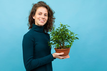 Young And Beautiful Caucasian Girl With Curly Hair Keeps Green Flower In A Pot And Smiles, Portrait Isolated On Blue Background