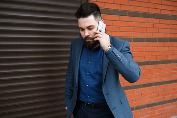 Wall Mural - Stylish handsome brunette man with beard, wearing classic formal blue suit jacket, talking on a smartphone outdoors on the city street near garage roller door. Businessman using mobile phone.