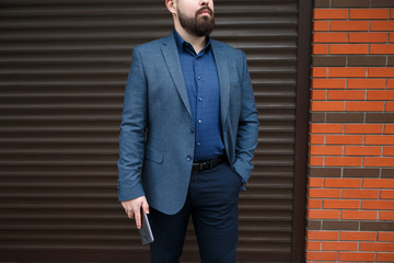 Wall Mural - Stylish handsome brunette man with beard, wearing blue suit, standing with smartphone in his hands against street wall. Classic elegant formal men's outfit.