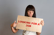 © _KUBE_ - Business and advertising. A young brunette with a piercing holding a cardboard with text LOOKING FOR A JOB and points a finger at it. Gray background