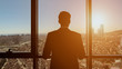 Silhouette of successful young businessman in suit is looking at window. He admires panoramic city view from his modern apartment, back view.