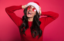 Crazy Party. Wonderful Girl In A Red Knitted Sweater And Pink Sunglasses Is Posing With Her Hands On Her Head, Holding Her Santa Claus Hat And Laughing With Her Eyes Closed.
