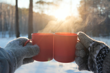 young man and young woman in knitted wool mittens with mugs of hot drink on Sunny frosty day on forest background