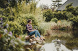 © Westend61 - Woman sitting at garden pond, using digital tablet
