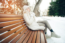 Young Beautiful Blonde Girl Sits On A Bench In The Park In Winter